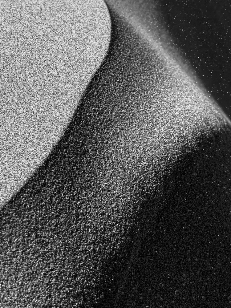 Black-and-white abstract of wind-swept sand dunes with the sun low in the sky, creating dramatic tonal contrast and tiny white specks of blowing sand across the image.