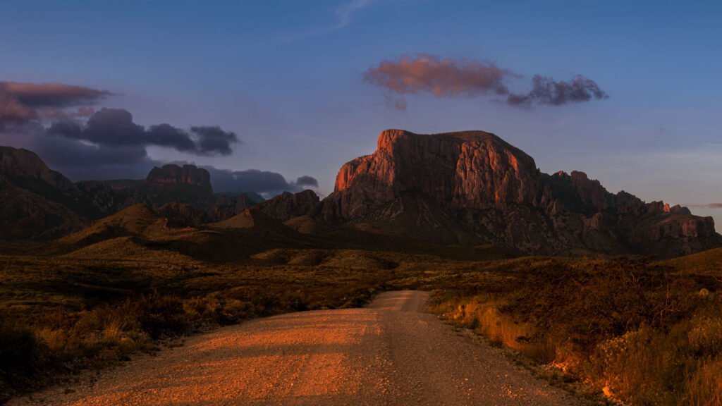 Gravel road leading toward the Chisos Mountains in Big Bend National Park, with warm late-day sunlight illuminating the road and mountain faces beneath a blue sky.