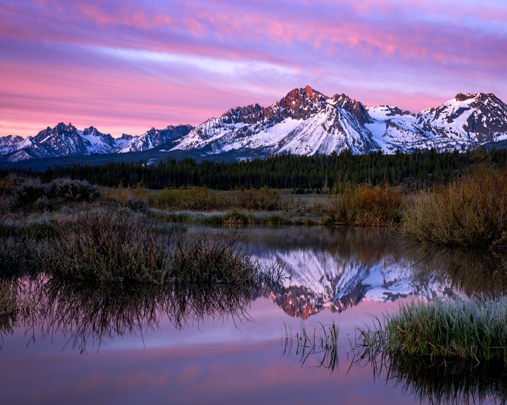 Mount Heyburn in Idaho’s Sawtooth Mountains at sunrise, glowing with alpenglow and reflected in the calm water below beneath a pink and purple sky.