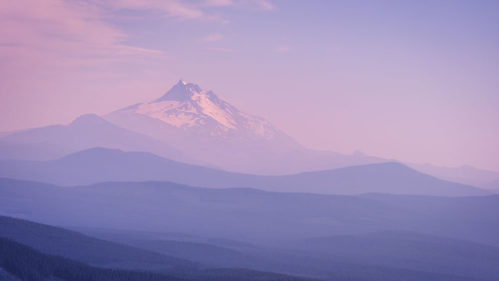 Mount Thielsen in Oregon rising above layered mountain ridges, softened by mist and wildfire haze beneath a pale pink morning sky.