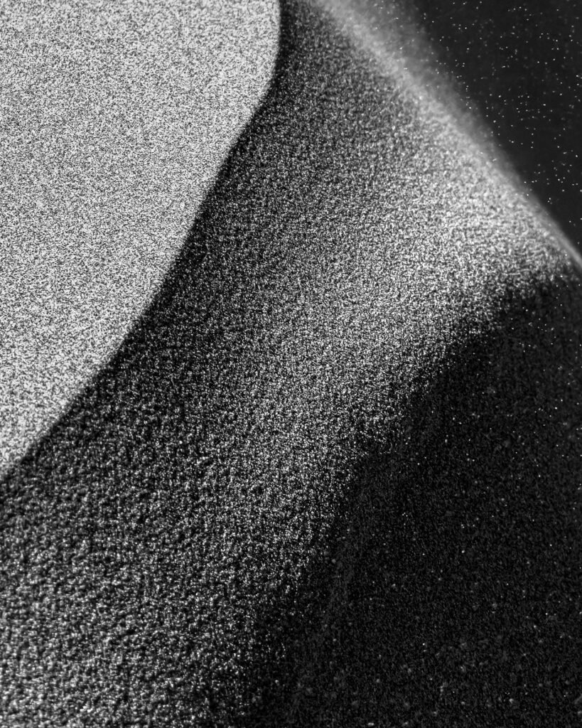 Black-and-white abstract of wind-swept sand dunes with the sun low in the sky, creating dramatic tonal
contrast and tiny white specks of blowing sand across the image.