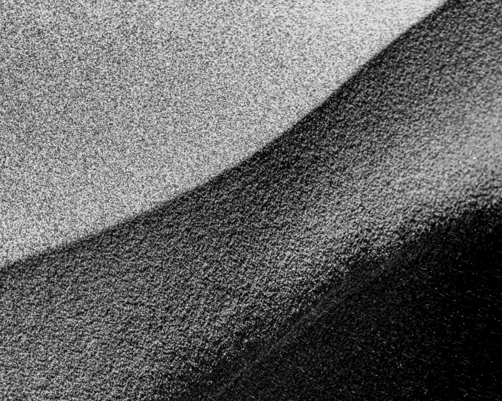 Black-and-white abstract of wind-swept sand dunes with the sun low in the sky, creating dramatic tonal contrast and tiny white specks of blowing sand across the image.