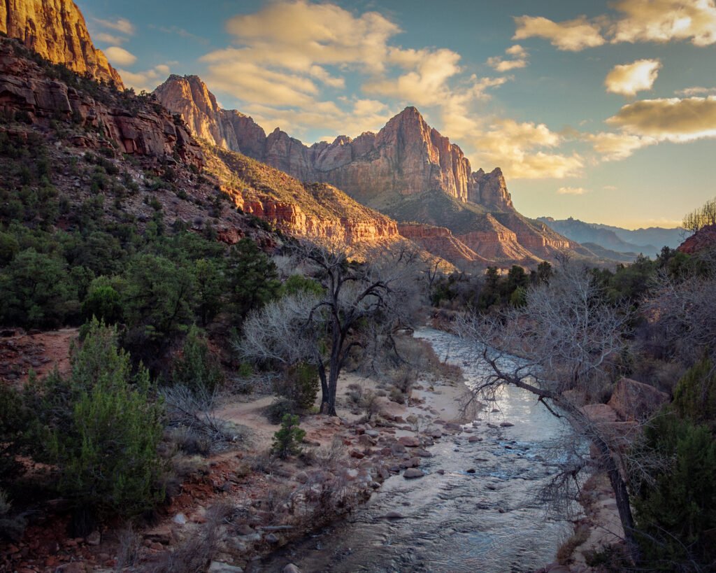 The Watchman rises above the Virgin River in Zion National Park, Utah. Photographed from a bridge shortly before sunset, warm golden light illuminates the red sandstone cliffs and scattered clouds, while trees and the winding river fill the shadowed foreground.